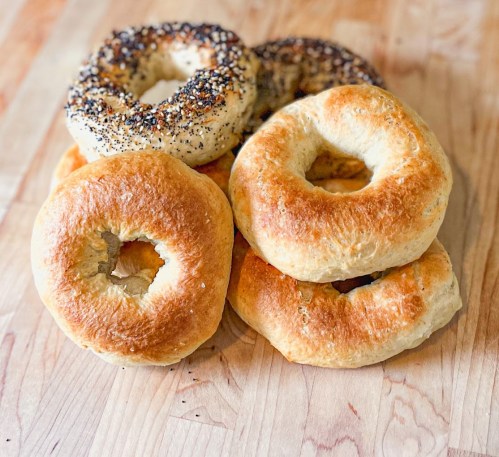 A stack of freshly baked bagels, including plain and everything bagels, on a wooden surface.