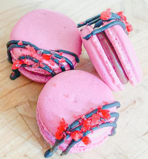 Three pink macarons drizzled with chocolate and topped with red sugar crystals, arranged on a wooden surface.