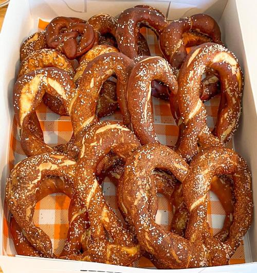 A box filled with various freshly baked pretzels, some sprinkled with salt, arranged neatly on an orange checkered paper.