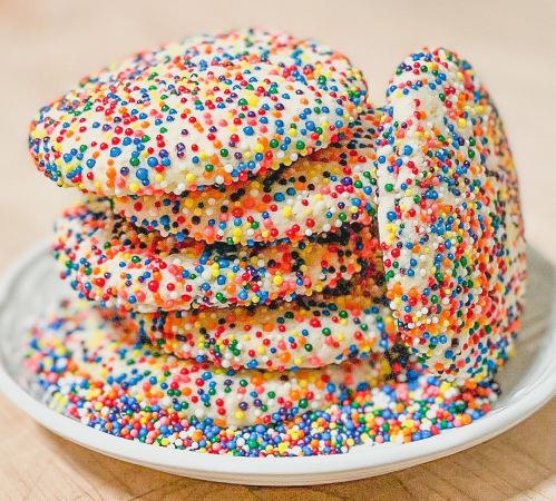 A plate stacked with colorful, sprinkle-covered cookies, surrounded by scattered sprinkles on the surface.