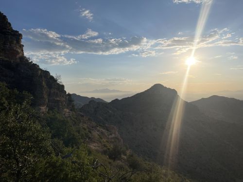 Sunset over rugged mountains, with a warm glow illuminating the landscape and clouds in the sky.