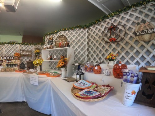 A cozy market display with seasonal decorations, snacks, and crafts on white tables against a lattice backdrop.