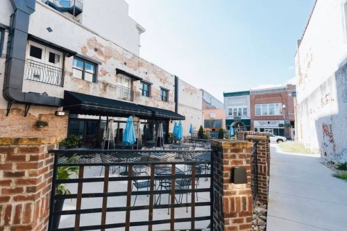 Outdoor dining area with blue umbrellas, brick walls, and a view of nearby buildings under a clear blue sky.
