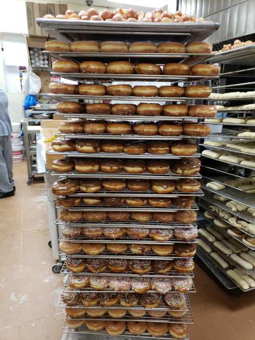 A tall stack of trays filled with various donuts in a bakery, with a person working in the background.