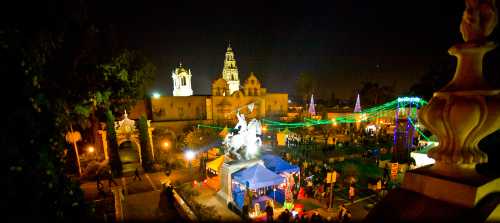 A vibrant night scene of a park with festive lights, tents, and a statue, surrounded by historic buildings.