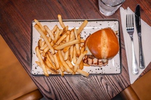 A plate with a grilled chicken sandwich on a bun and a side of golden French fries, set on a wooden table.