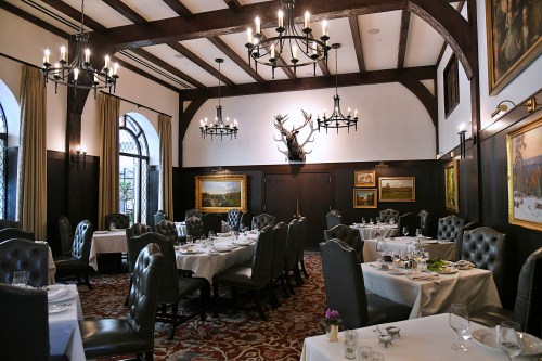 Elegant dining room with dark wood beams, chandeliers, and tables set for dinner, featuring a mounted deer head on the wall.
