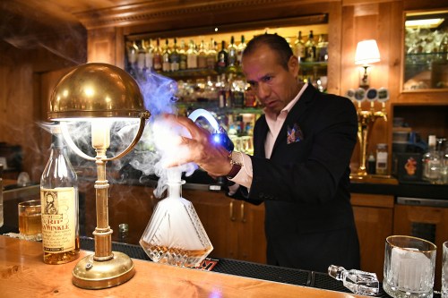 A bartender mixes a drink with smoke effects, surrounded by bottles in a stylish bar setting.