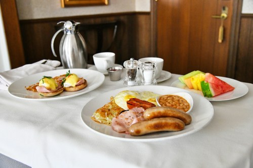 A breakfast spread featuring eggs Benedict, sausages, baked beans, and fresh fruit on a table.