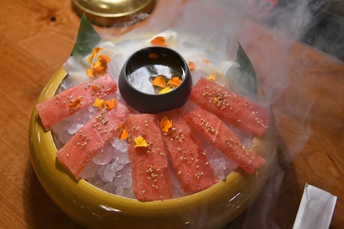 A vibrant dish featuring sliced tuna on ice, garnished with petals and sesame seeds, with a small bowl of sauce.
