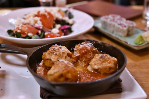 A cast iron skillet with meatballs topped with cheese, alongside a salad and sushi rolls on a wooden table.
