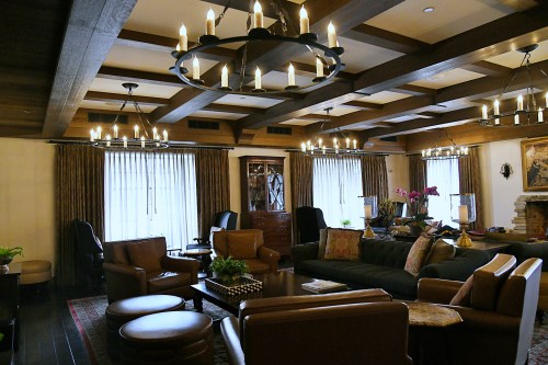Cozy living room with wooden beams, chandeliers, comfortable seating, and large windows adorned with curtains.