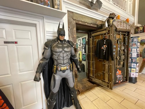 A life-sized Batman statue stands near a door in a shop, surrounded by various merchandise and decorations.