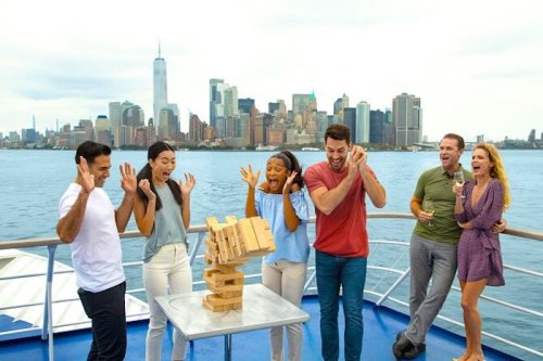A group of excited people on a boat play Jenga with a city skyline in the background, expressing surprise and joy.
