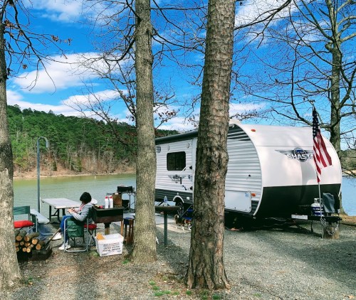A camper by a lake, with a person sitting at a picnic table, surrounded by trees and a clear blue sky.