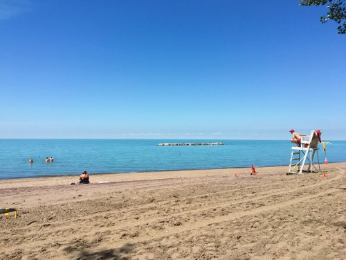 A sandy beach with calm water, a lifeguard chair, and a few people swimming under a clear blue sky.
