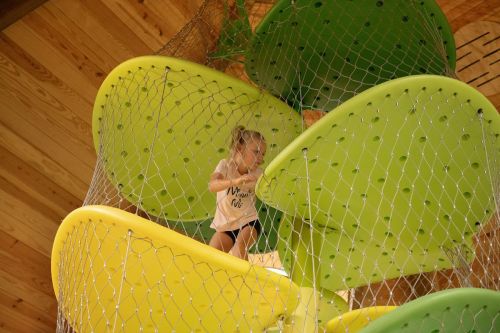 A child climbs on colorful, curved play structures surrounded by safety netting in a wooden play area.