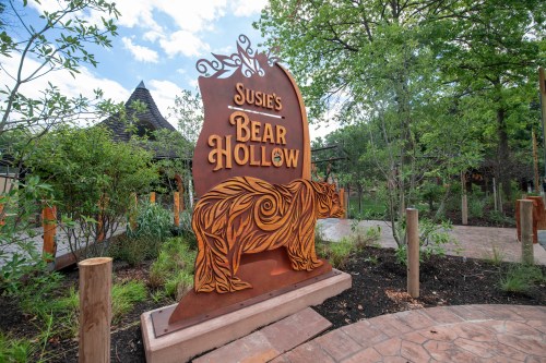 Sign for "Susie's Bear Hollow" featuring a carved bear design, surrounded by greenery and a pathway.