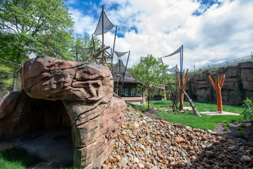 A playground with rock formations, wooden structures, and trees, surrounded by greenery and a cloudy sky.