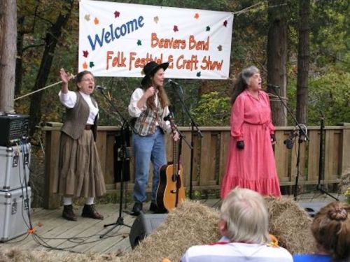 Three performers sing on stage at a folk festival, with a welcome banner in the background and an audience in front.