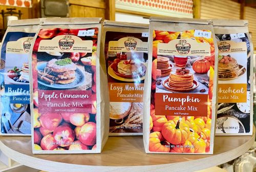 Five varieties of pancake mix bags displayed on a wooden table, featuring flavors like apple cinnamon and pumpkin.
