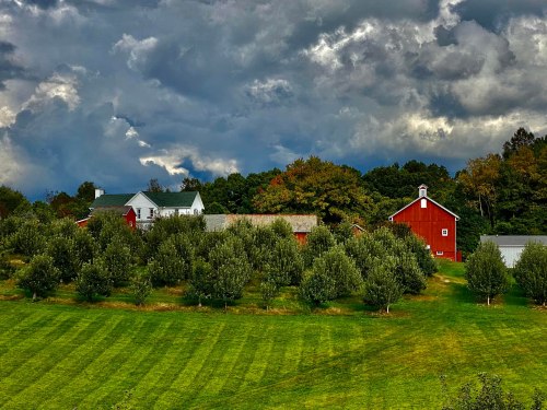 A scenic farm with red barns and a house, surrounded by green fields and trees under a dramatic cloudy sky.