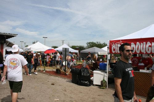 A busy outdoor market scene with tents, people walking, and food stalls under a partly cloudy sky.