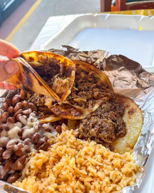 A hand holds a taco filled with meat, alongside rice and beans on a foil-lined plate.