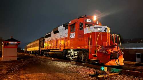 A bright orange train engine with the number 501 parked on tracks at night, with a vintage passenger car behind it.