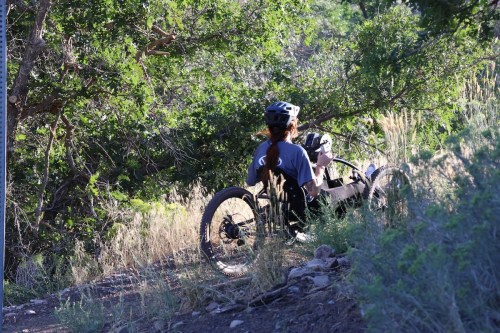 A person in a helmet sits on a handcycle on a dirt path surrounded by greenery.