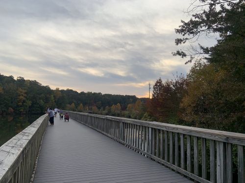 A wooden walkway stretches over a lake, with people walking and colorful autumn trees lining the shore under a cloudy sky.
