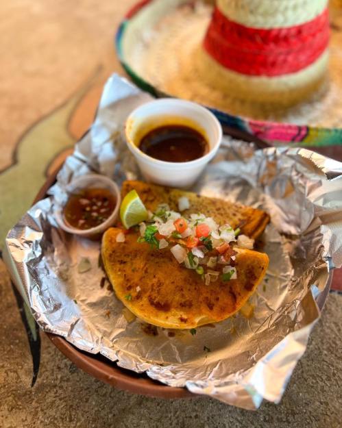 A taco on foil with toppings, accompanied by two small cups of sauce and a lime wedge, with a sombrero in the background.