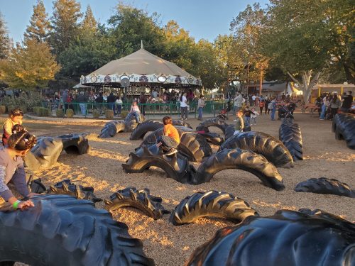 Children play on large black tire structures in a park, with a carousel and trees in the background.