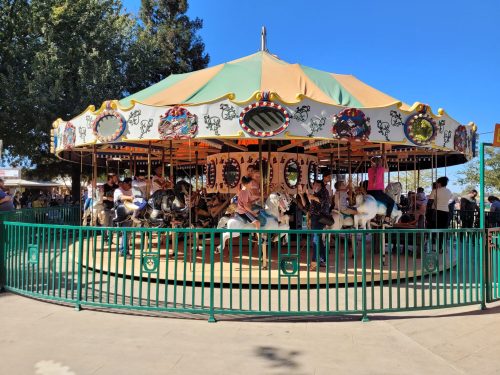 A colorful carousel with horses, surrounded by a green fence, and people enjoying a sunny day at an amusement park.