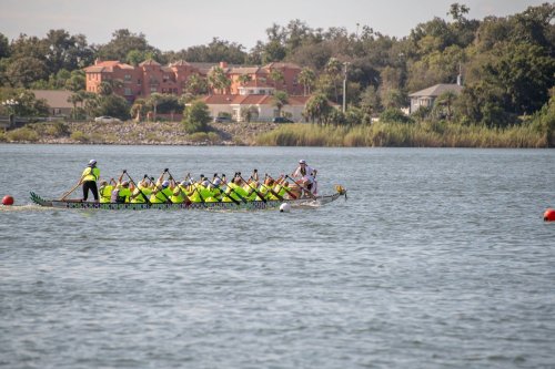 A dragon boat team paddles in a lake, with spectators and buildings visible in the background.