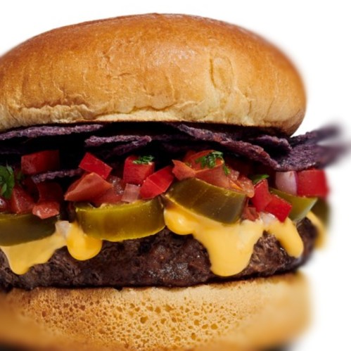 A close-up of a burger with a juicy beef patty, melted cheese, tomatoes, pickles, and blue corn tortilla chips.