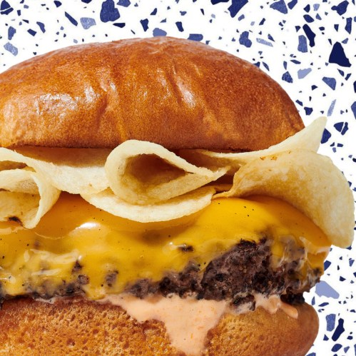 Close-up of a burger with melted cheese, crispy potato chips, and a creamy sauce, set against a colorful background.