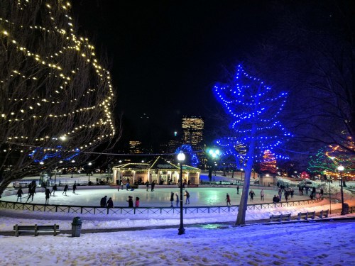 A winter scene of an ice skating rink at night, surrounded by colorful lights and snow-covered trees.