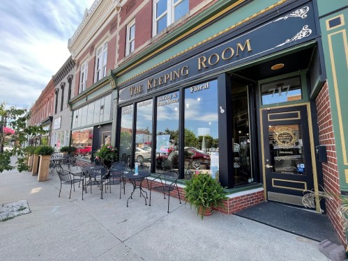 Exterior of "The Keeping Room" shop with outdoor seating and floral displays on a sunny day.