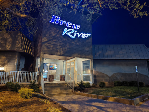 A modern building at night with a glowing "Brew River" sign and a welcoming entrance surrounded by greenery.