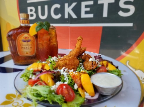 A vibrant salad with fried chicken, mango, tomatoes, and a dipping sauce, alongside a cocktail and a bottle of peach whiskey.