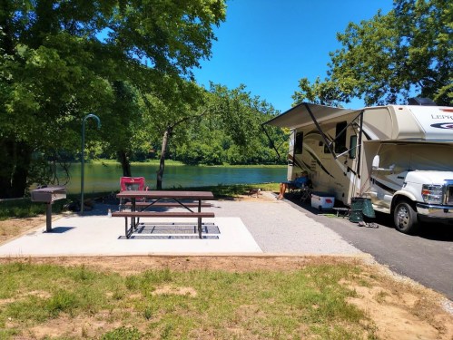 A sunny campsite by a river featuring an RV, picnic table, grill, and lush green trees.