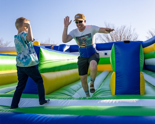 A man in a cap playfully jumps on a colorful inflatable while a child watches, both enjoying a sunny day outdoors.