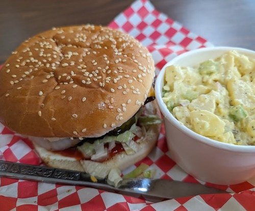 A sesame seed bun sandwich with lettuce and ketchup, served alongside a cup of creamy potato salad.