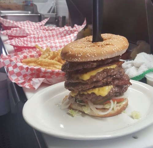 A towering burger with multiple patties, cheese, lettuce, and tomato, served on a plate with fries in the background.