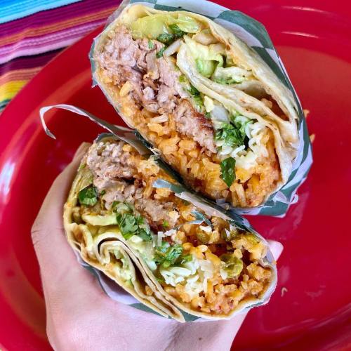A hand holding two burritos, one with beans and rice, the other with lettuce and cilantro, on a red plate.