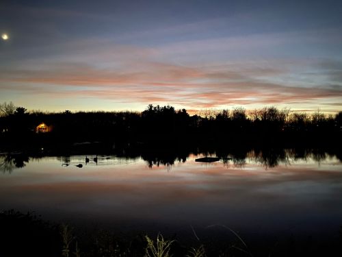 A serene lake at dusk, reflecting colorful clouds and silhouettes of trees, with a hint of moonlight above.