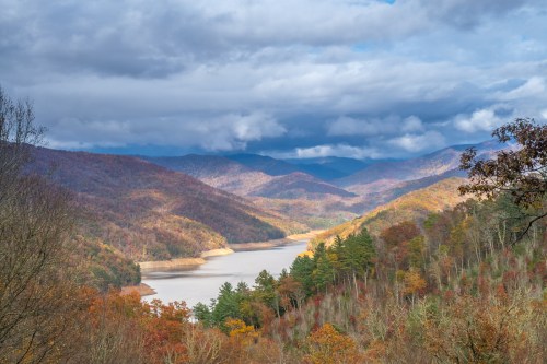 A scenic view of a lake surrounded by mountains, with autumn foliage and dramatic clouds in the sky.