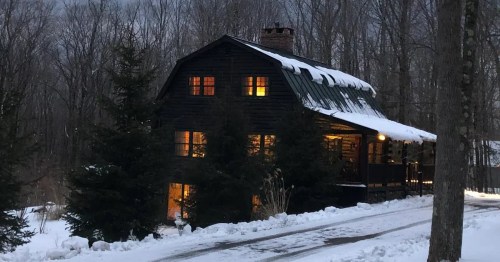 A cozy black cabin with warm lights, surrounded by snow and bare trees under a cloudy sky.