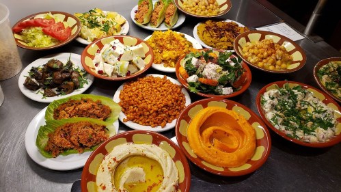 A variety of colorful dishes including salads, tacos, hummus, and rice, arranged on plates on a table.